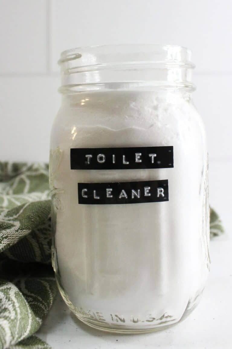 A glass jar labeled "Toilet Cleaner," filled with a white powder mix, sits beside a green and white patterned cloth. This easy homemade toilet bowl cleaner promises to bring sparkle and freshness to your bathroom routine.