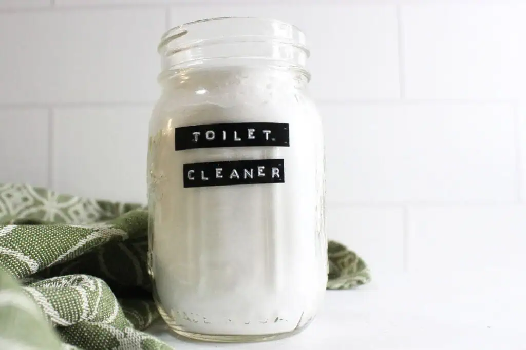 A glass jar filled with white powder, boldly labeled "Toilet Cleaner," sits elegantly next to a vibrant green patterned cloth.
