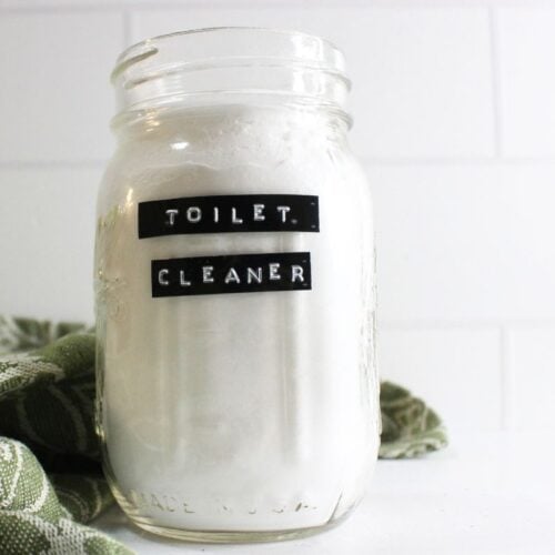 A glass jar filled with white powder, boldly labeled "Toilet Cleaner," sits elegantly next to a vibrant green patterned cloth.