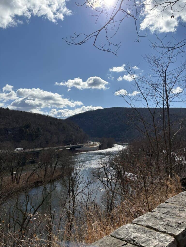 A river winds through a valley surrounded by hills and bare trees, with the sun peeking through a partly cloudy sky. In the background, a bridge gracefully spans the river. This serene scene invites personal reflection and authentic storytelling, capturing the essence of nature's beauty.