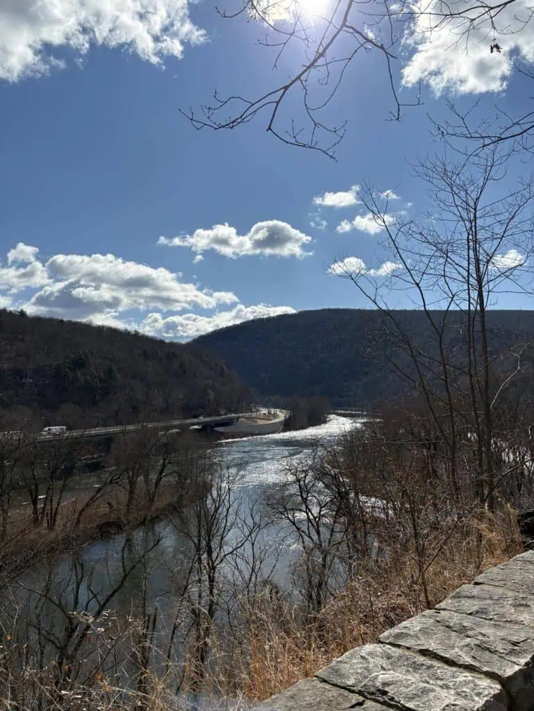 A river winds through a valley surrounded by hills and bare trees, with the sun peeking through a partly cloudy sky. In the background, a bridge gracefully spans the river. This serene scene invites personal reflection and authentic storytelling, capturing the essence of nature's beauty.