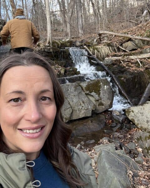 A woman captures a selfie in front of a tranquil forest waterfall, while in the background, someone in a brown jacket navigates uphill. It's a serene snapshot perfect for her personal blog.