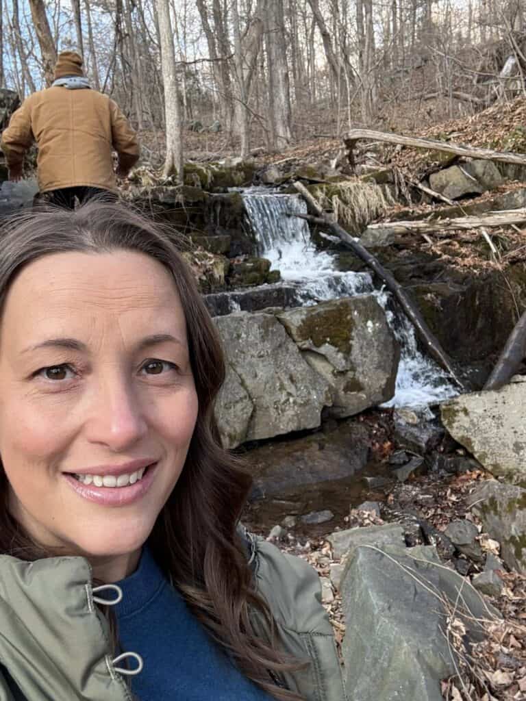 A woman captures a selfie in front of a tranquil forest waterfall, while in the background, someone in a brown jacket navigates uphill. It's a serene snapshot perfect for her personal blog.