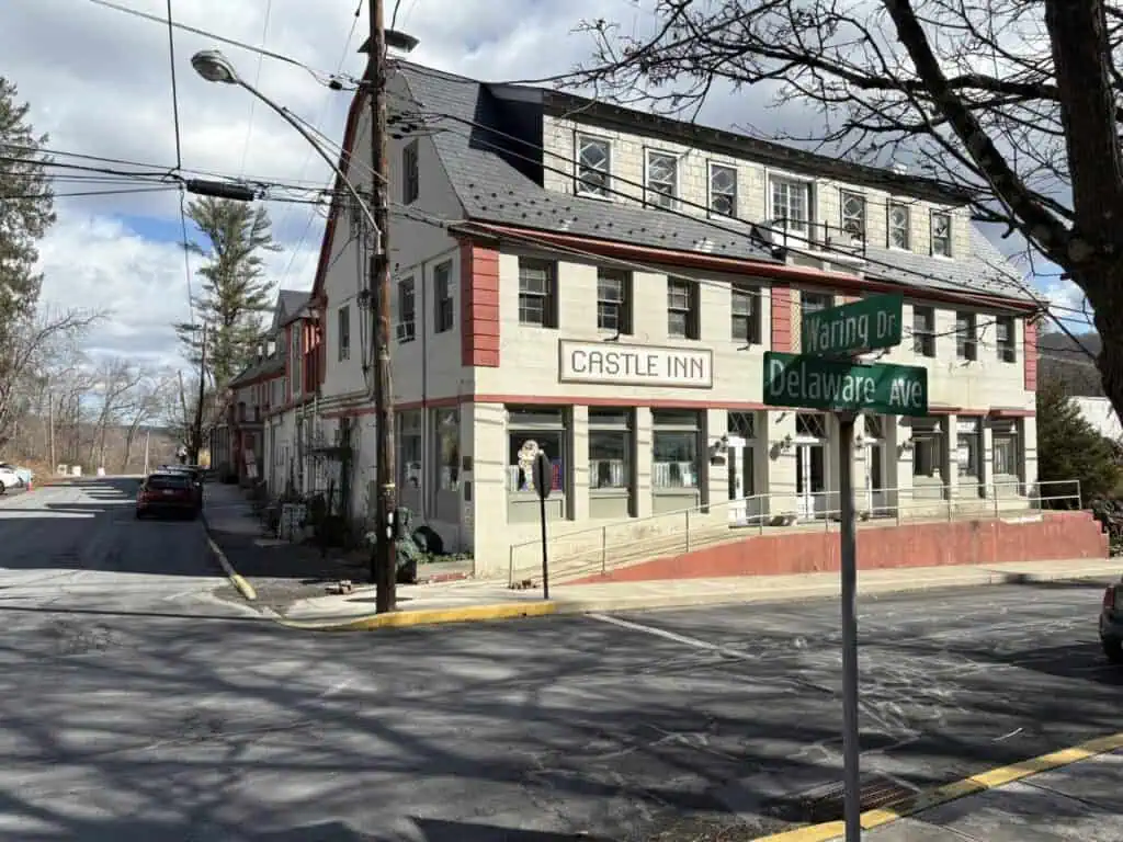 Street corner with a three-story building named Castle Inn. The intersection sign reads Warring Dr and Delaware Ave. Cars are parked along the street. Cloudy sky in the background.