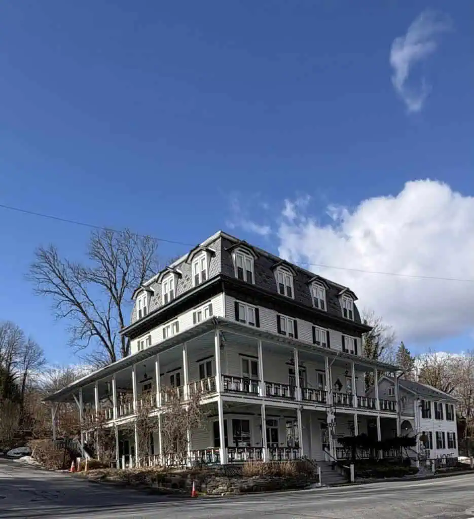 A large, multi-story white building with a wraparound porch stands at a street corner. The sky is clear, with a few clouds. Leafless trees surround the area.