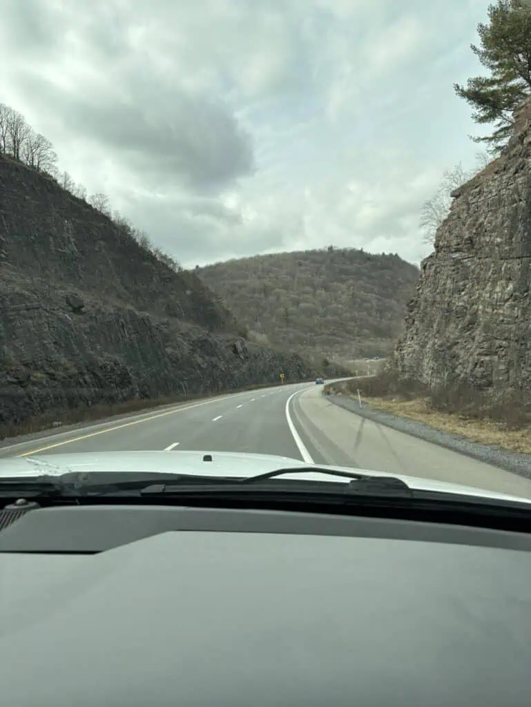 Personal perspective from the car dashboard reveals a road gracefully curving through rocky hills under a cloudy sky. Sparse vegetation dots the hillsides, evoking a sense of serene isolation, as if in a postcard scene.