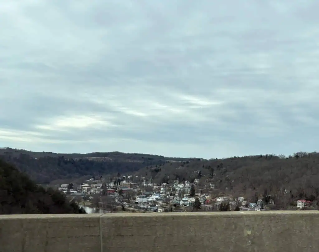 View of a small town nestled in a valley, surrounded by rolling hills and under a cloudy sky, as seen from a concrete barrier.
