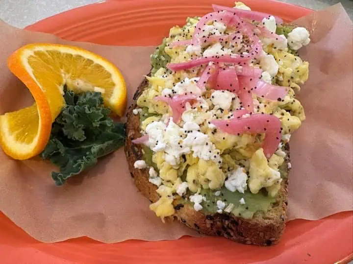 Avocado toast topped with scrambled eggs, pickled onions, and cheese on a plate, garnished with orange slices and kale.