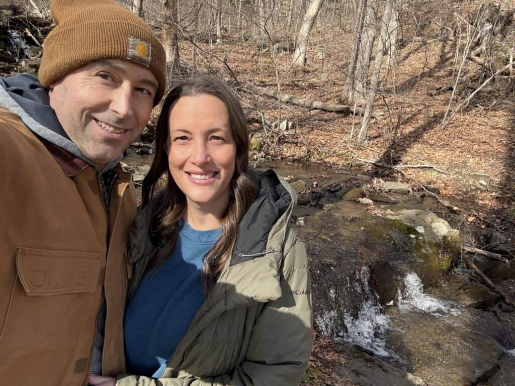 A man and woman stand in front of a small waterfall in a forest during autumn, both wearing jackets and posing for a personal post as they look at the camera.