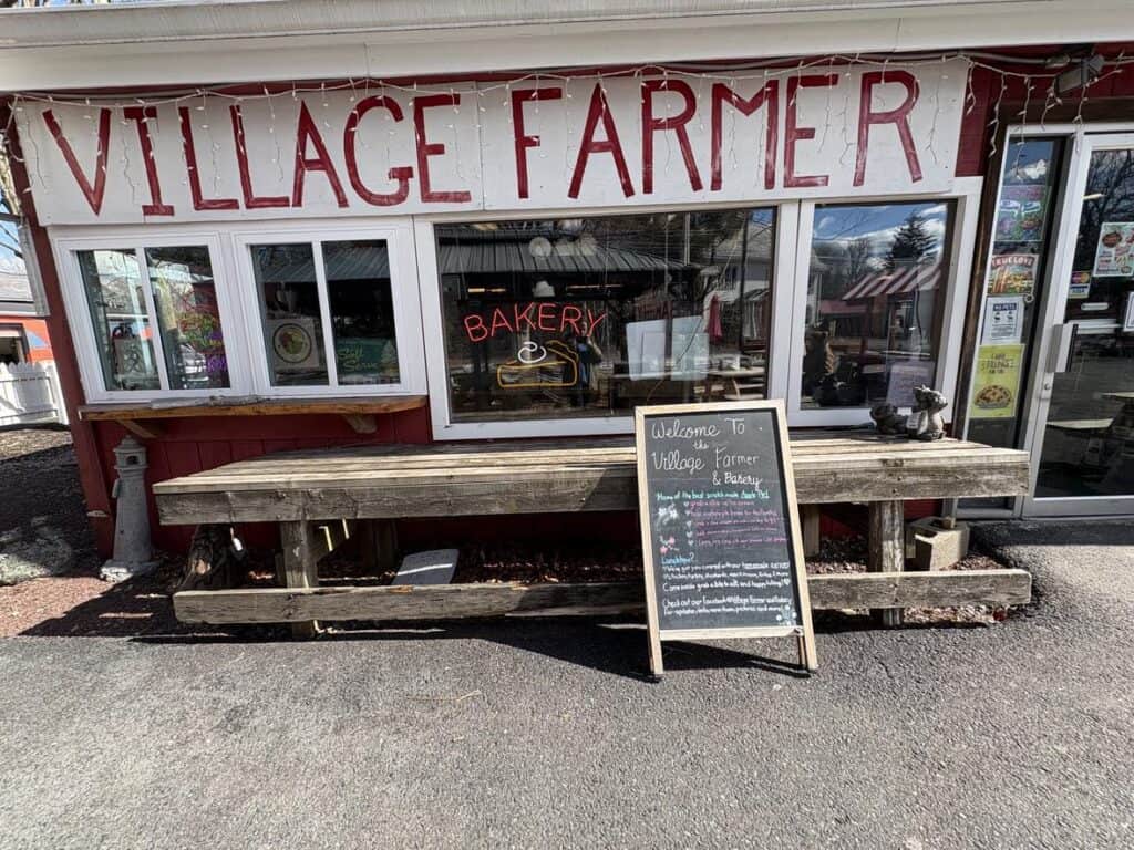 The exterior of Village Farmer and Bakery exudes a personal touch with a wooden bench and chalkboard menu inviting passersby. A neon "Bakery" sign in the window adds charm, reminiscent of the perfect spot to post up and enjoy the day.