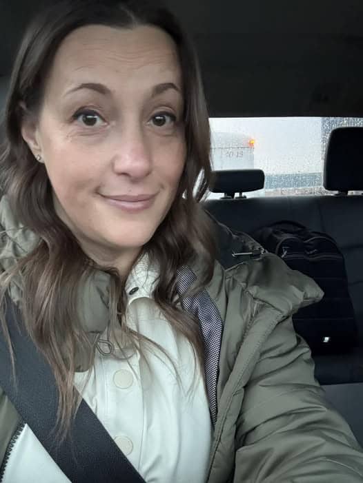 A person with brown hair, dressed in a personal green jacket, smiles slightly while wearing a seatbelt inside a car post-drive.