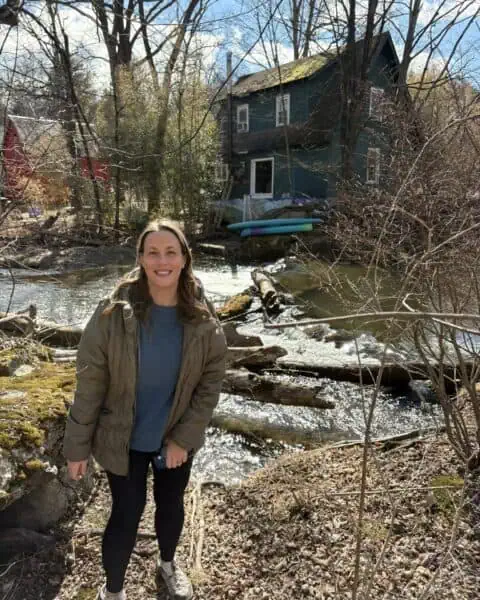 Woman stands by a creek with fallen logs and trees, in front of a green house. She is wearing a jacket and holding a drink bottle. It's a sunny day with clear skies.