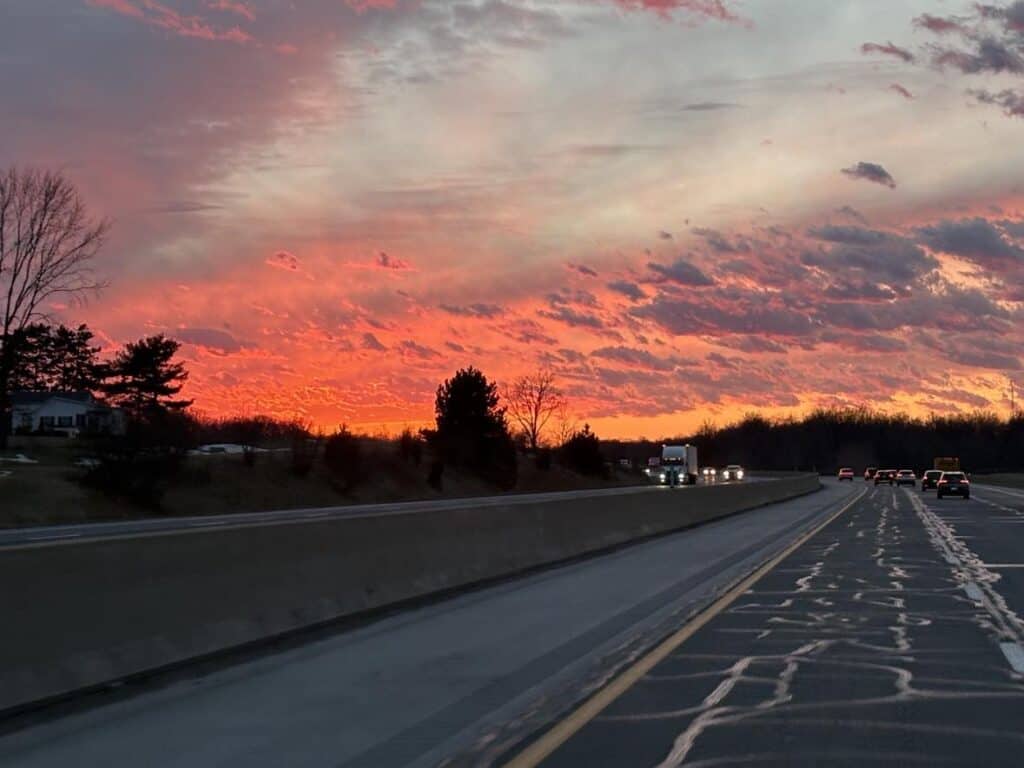 A highway at dusk, where cars post their personal journeys in both directions. The sky is a vibrant mix of pink, orange, and purple hues with scattered clouds. Trees line the road on the left side, adding to the serene landscape.