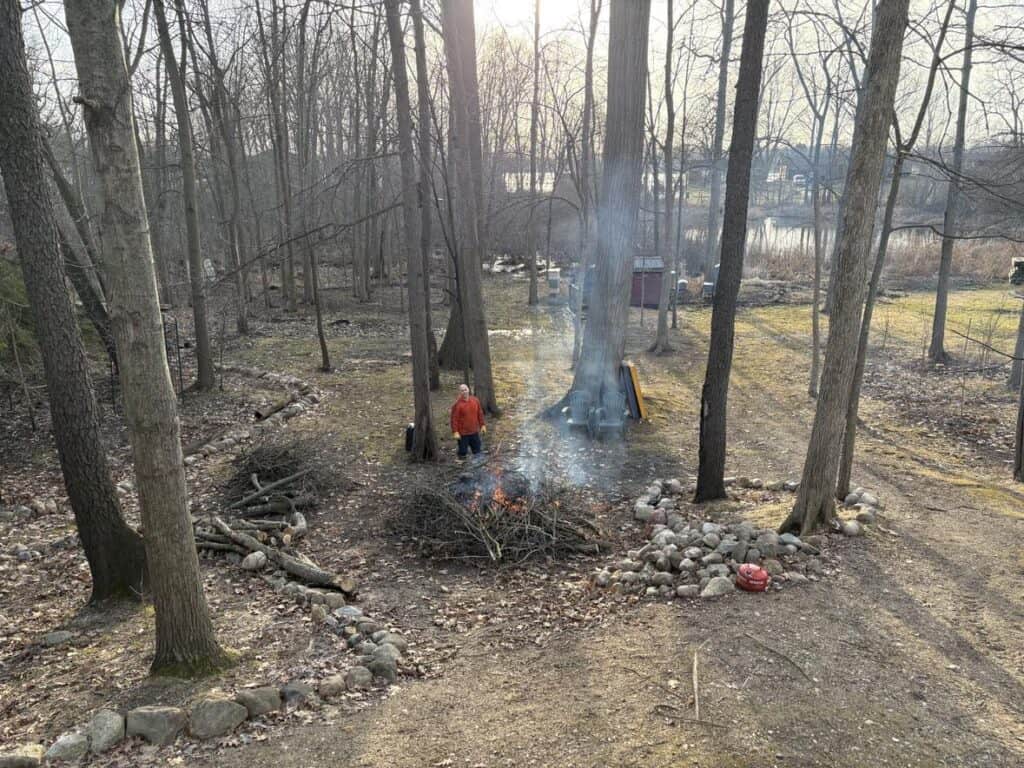 A person stands near a small, smoking bonfire in a wooded area, finding a personal moment of reflection surrounded by scattered fallen branches. In the background, the tranquil lake post-sunset offers a serene view.