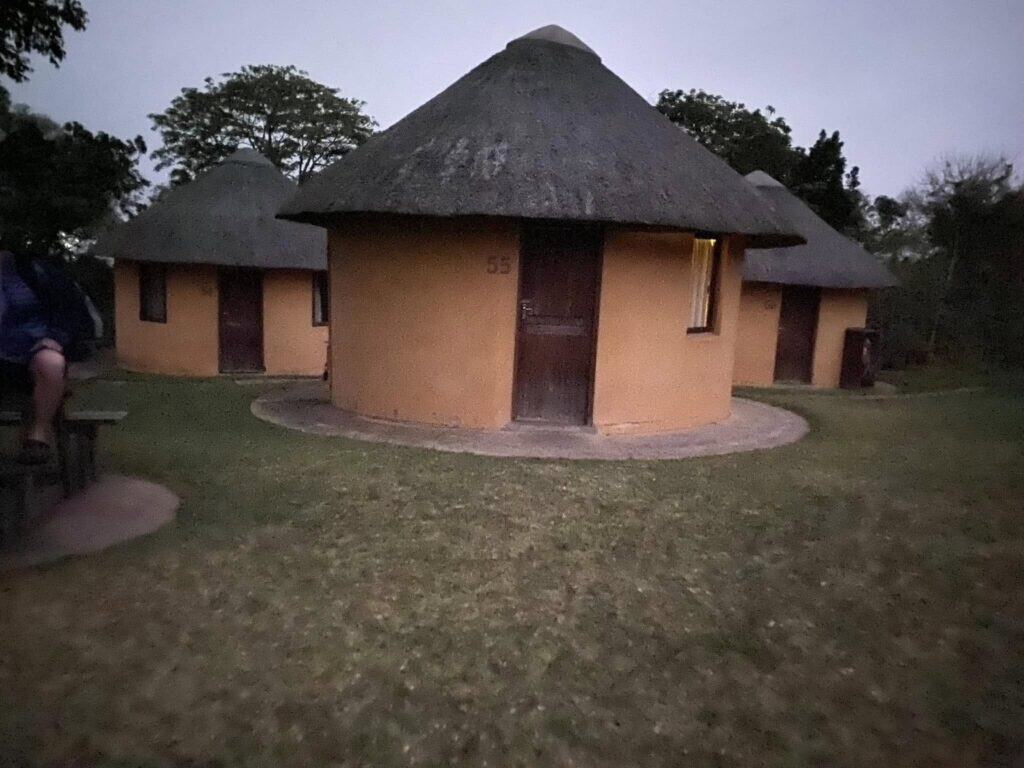 At dusk, three round huts with thatched roofs and wooden doors sit in a grassy area, framed by trees in the background. The scene evokes a sense of personal tranquility, like stepping into a serene post-card from nature.