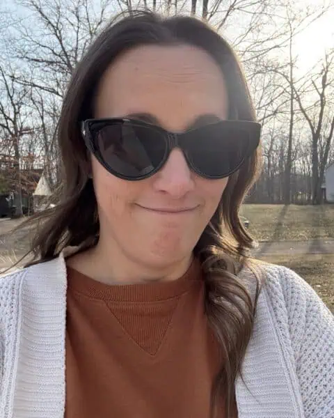 A person wearing chic sunglasses and a white cardigan stands outside amidst leafless trees, basking in the golden sunlight.
