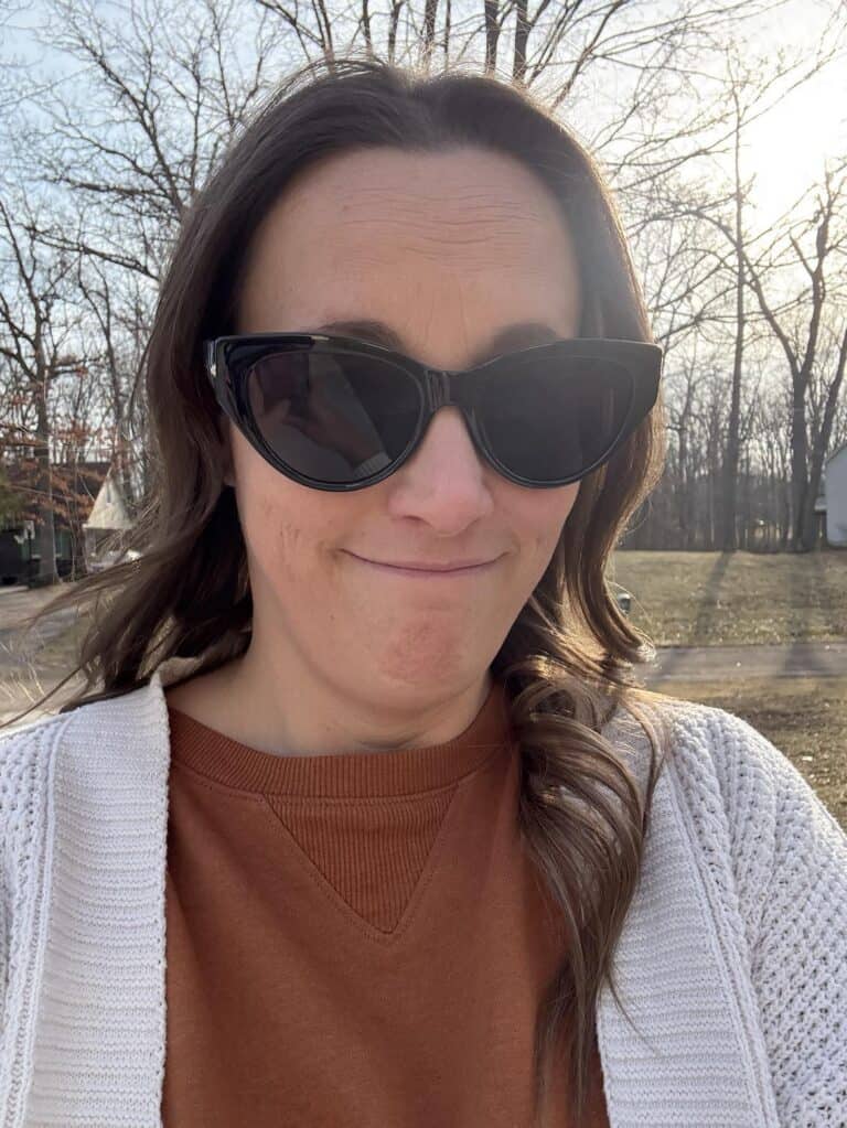 A person wearing chic sunglasses and a white cardigan stands outside amidst leafless trees, basking in the golden sunlight.