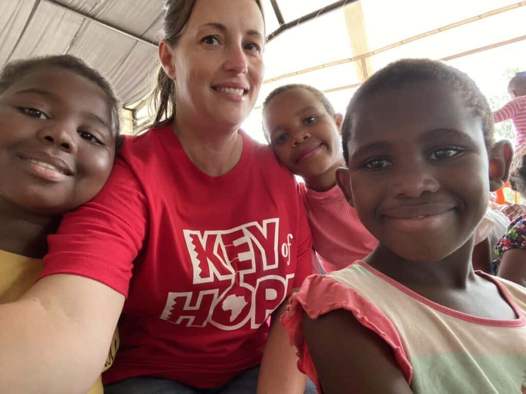 A person in a red "Key of Hope" shirt takes a selfie with three children beaming around them, sitting under a tent—a moment that feels like a fresh start.