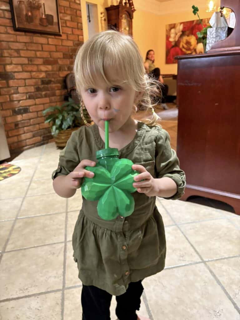 In a cozy personal post, a young child sips from a green clover-shaped container with a straw, standing indoors against the backdrop of a brick wall and wooden cabinet.