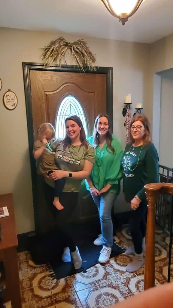 Four people stand indoors near a door, each with a personal connection that radiates warmth. One person holds a small child, and they're all smiling, dressed casually as if posing for a cherished post.