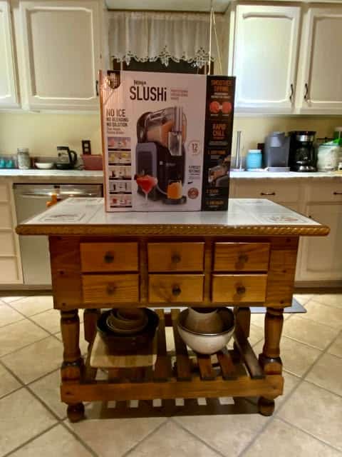 A wooden kitchen island with drawers holds a boxed Nova Slushi machine, perfect for crafting icy treats. Kitchen appliances and cabinets create a warm backdrop, ideal for sharing on a personal blog or social media content.