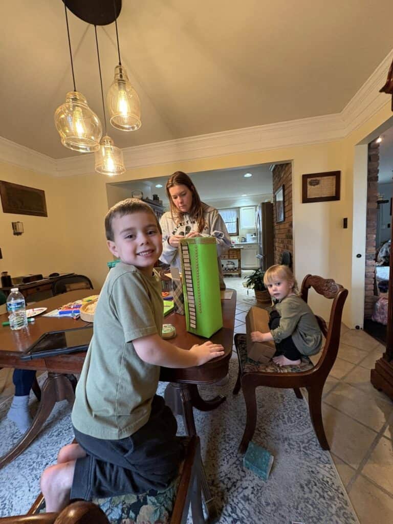 Three children are gathered around a wooden table. One is standing, smiling for a personal photo, while another sits on a chair. The third is focused on a tall stack of green folders, perhaps crafting the perfect post for later.