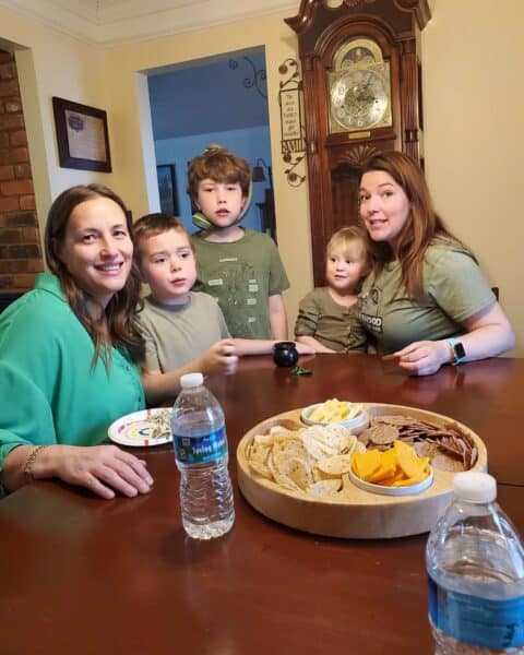 Two women and three children gather around a cozy table, enjoying snacks and refreshing bottled water. In the background, a wall clock quietly ticks away, marking their precious moments together.