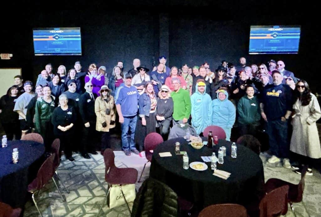 A lively group gathers in a spacious room with round tables, many donning playful or themed costumes. Behind, two screens glow softly, capturing the festive spirit of this memorable personal post.