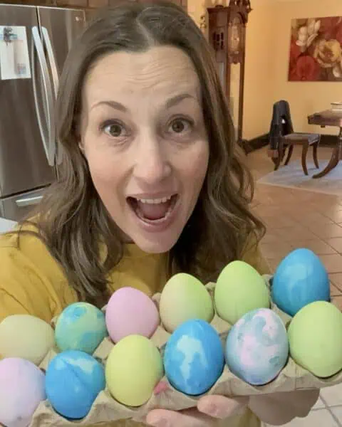 In a personal post, a woman smiles with delight, proudly displaying a carton of colorful dyed eggs in her cozy kitchen setting.