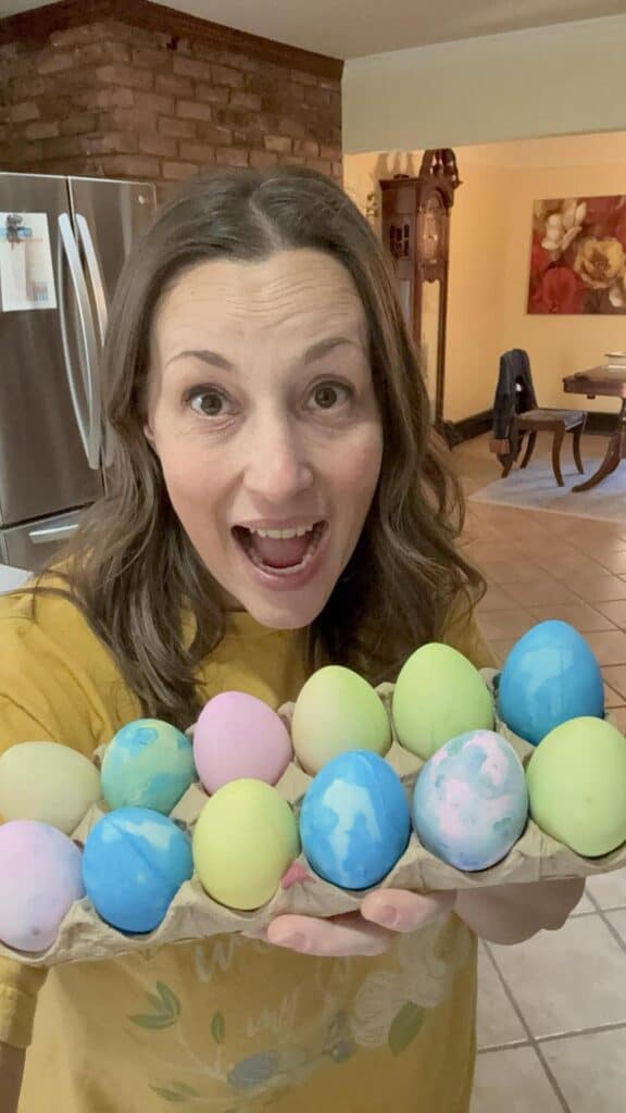 In a personal post, a woman smiles with delight, proudly displaying a carton of colorful dyed eggs in her cozy kitchen setting.