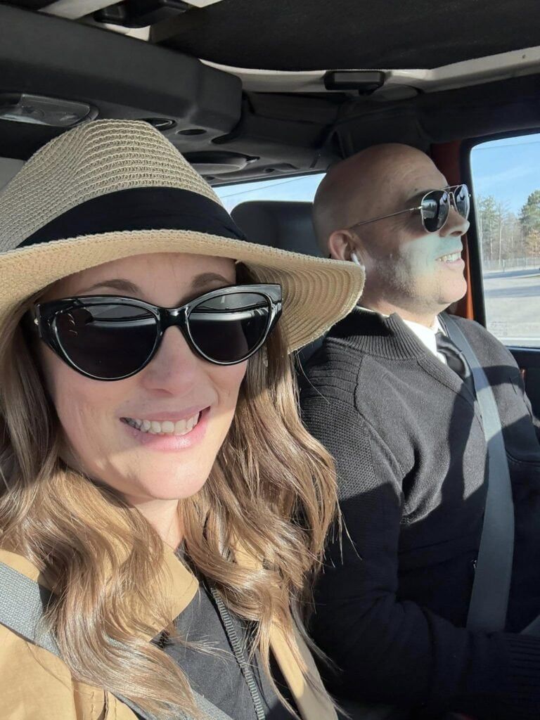 A woman in a straw hat and sunglasses sits next to a man, both smiling, as they snap a personal photo to post later.
