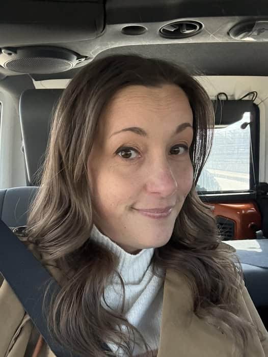 In a personal post, a woman with long brown hair and a beige coat sits in a car, wearing her seatbelt. She exudes warmth with her slight smile and cozy white turtleneck sweater.