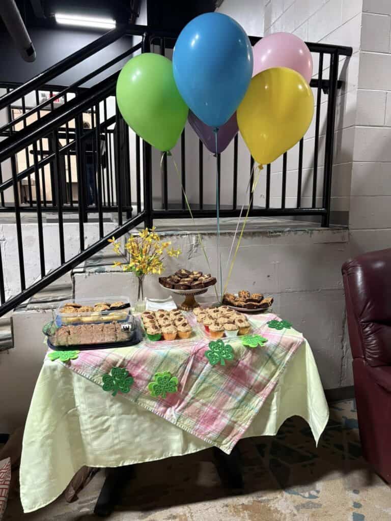 A table with assorted pastries and brownies sits under five colorful balloons, covered with a green and pink cloth. A small vase with yellow flowers adds a personal touch to the setting, reminiscent of a delightful post celebrating life’s little moments.