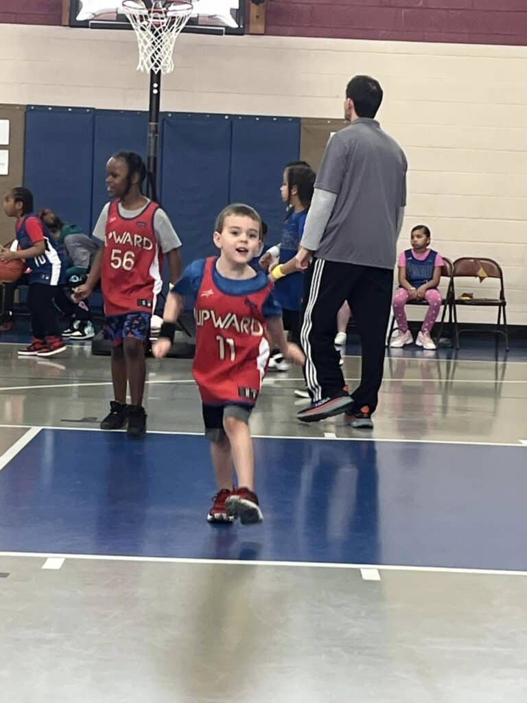 A young child in a red sports jersey joyfully runs across an indoor basketball court, capturing a moment of playful energy. In the background, other children and an adult share the lively scene. #PersonalPost.