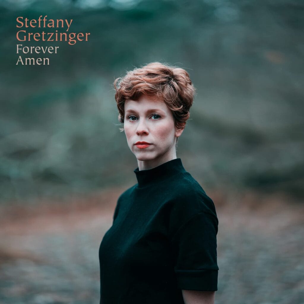 A person with short hair in a black shirt stands outdoors, embodying the soulful essence of Steffany Gretzinger's "Forever Amen." The serene backdrop perfectly frames a moment of contemplation and musical devotion.