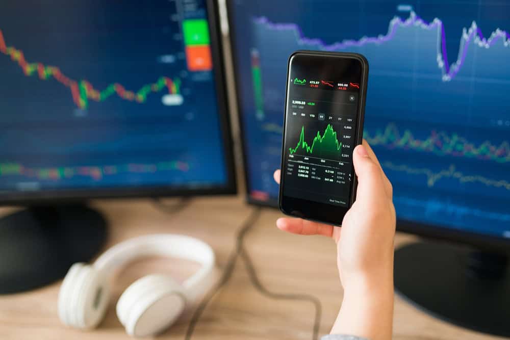 A person holds a smartphone displaying stock market charts in front of two monitors with similar graphs, diving deep into the world of investing. White headphones rest on the desk, ready to accompany their pursuit to make money through stocks.