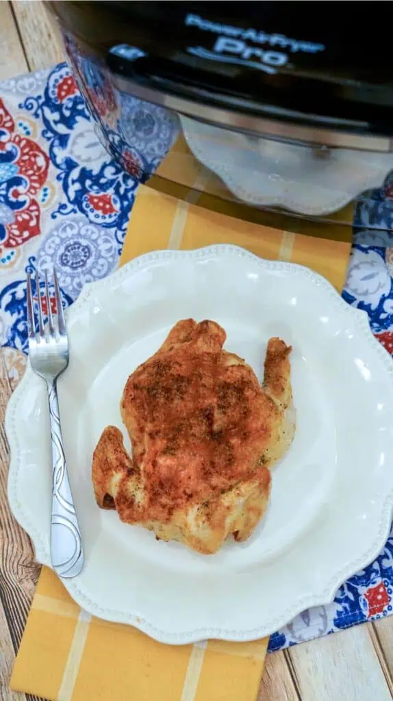 Roasted chicken on a white plate next to a patterned fork sits on a yellow placemat with a patterned tablecloth. An air fryer for that perfect roast cornish hen is tucked in the background, waiting for its next culinary adventure.