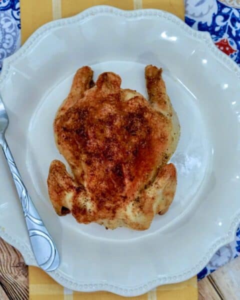 An air fryer Cornish hen sits roasted and golden on a white plate, accompanied by a fork, all set atop a vibrant patterned tablecloth.