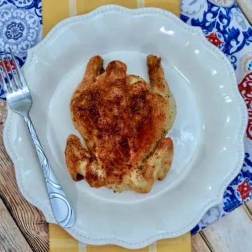 An air fryer Cornish hen sits roasted and golden on a white plate, accompanied by a fork, all set atop a vibrant patterned tablecloth.