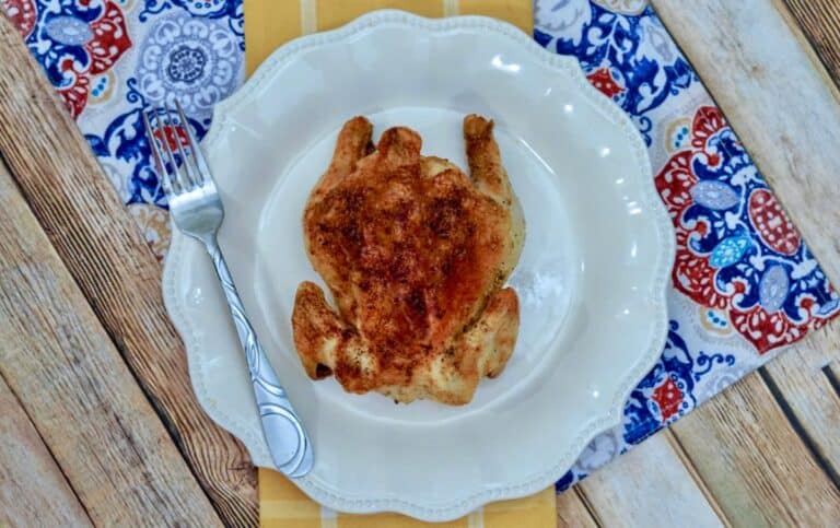 An air fryer Cornish hen sits roasted and golden on a white plate, accompanied by a fork, all set atop a vibrant patterned tablecloth.