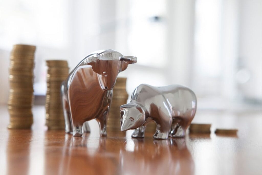 Silver bull and bear figurines rest on a wooden surface, symbolizing the thrilling world of investing, with stacks of coins in the background hinting at the potential to make money in stocks.