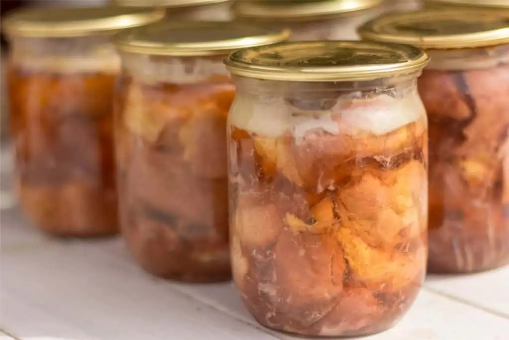 Several glass jars filled with preserved meat chunks and sealed with metal lids are arranged on a white wooden surface, showcasing an effective way to store meat without a refrigerator.