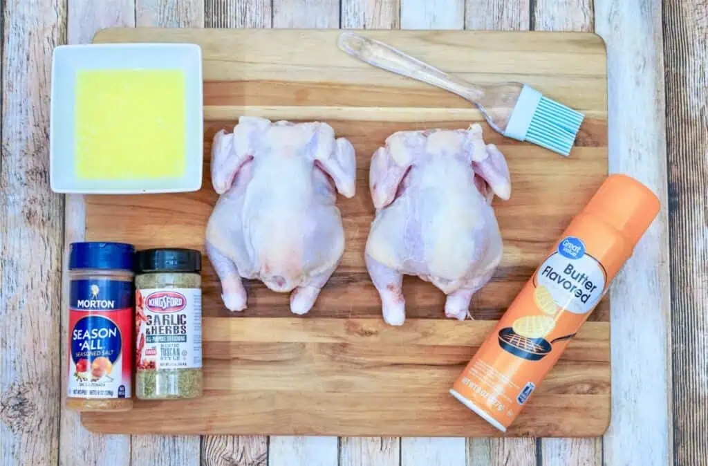 Two raw Cornish hens sit on a wooden cutting board alongside seasonings, melted butter, and a brush, ready to be transformed into a delicious air fryer Cornish hen meal. A quick spray of cooking oil sets the stage for perfectly crispy skin.