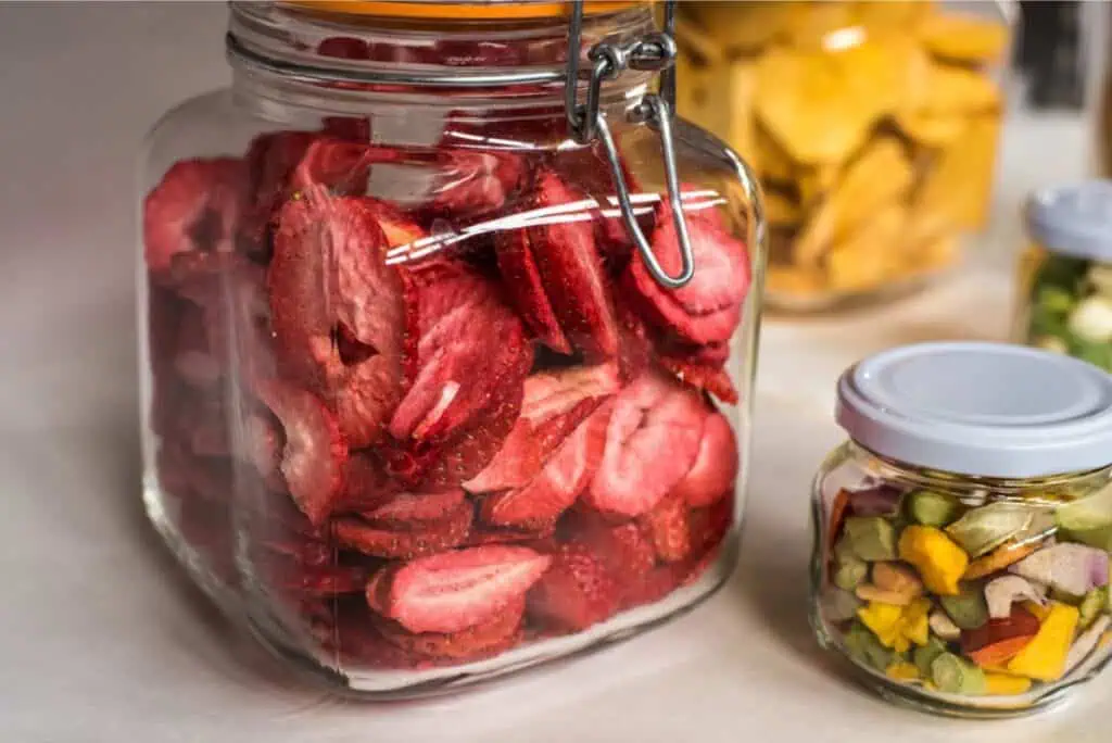 Glass jars filled with dried strawberry slices, a mix of dried fruits, and larger pieces of dried mango are displayed on a countertop, offering inspiration for those eager to get started with dehydrating food at home.
