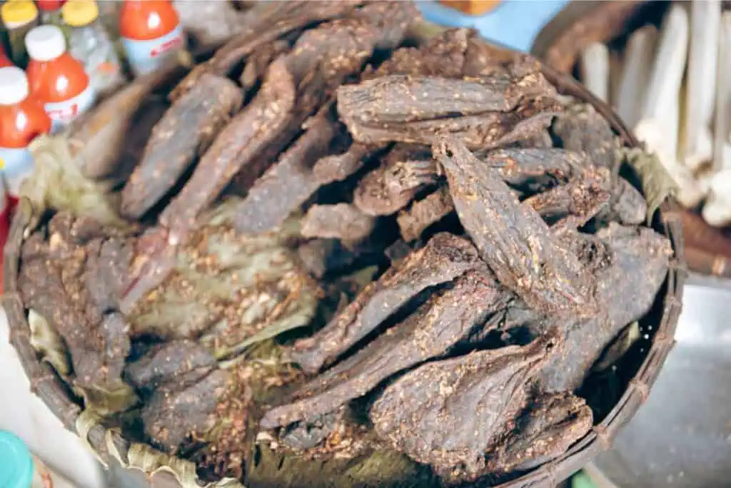 A basket filled with dark, dried meat slices is artfully displayed on a counter, showcasing traditional methods of meat preservation. In the background, bottles of red sauce and other goods add a splash of color to this charming display.