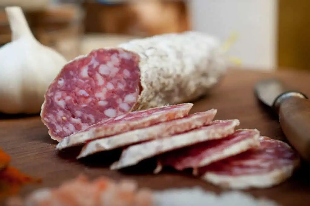 Sliced salami rests on a wooden cutting board, accompanied by a whole garlic bulb and a knife in the background, showcasing traditional meat preservation methods.