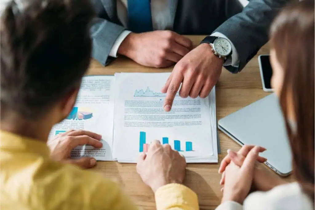 Three people sit at a table reviewing documents filled with charts and graphs. One person points at the papers, discussing retirement planning strategies, while a laptop nearby provides additional insights.