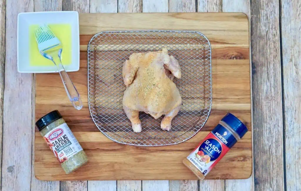A raw Cornish hen seasoned with spices sits on a wire rack, perched on a wooden cutting board. Nearby, a basting brush glistens with melted butter and two spice containers await. Perfect for an air fryer feast!.