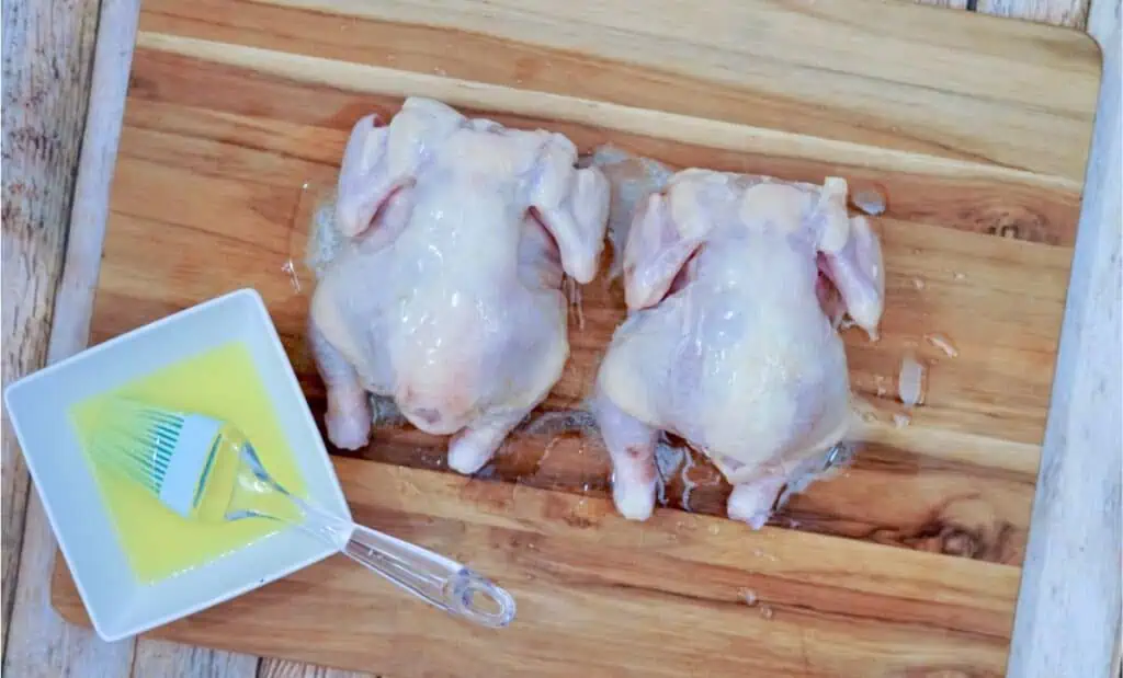 Two raw Cornish hens rest on a wooden cutting board, accompanied by a dish of melted butter and a basting brush, ready to transform into succulent delights when cooked in an air fryer.