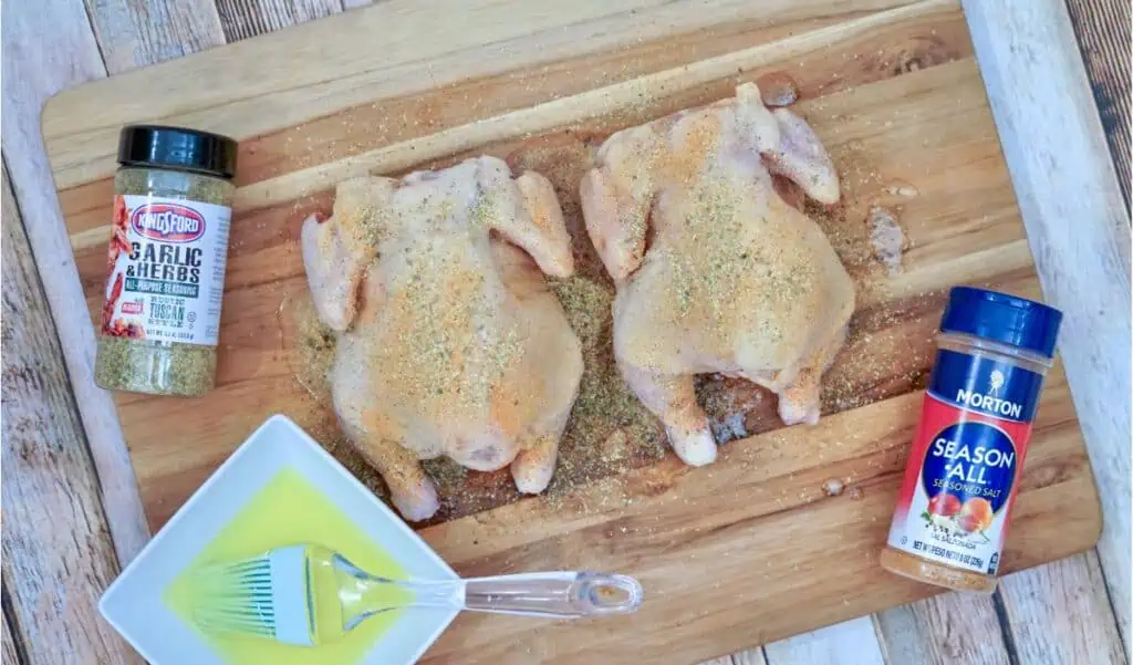Two seasoned raw chickens, reminiscent of preparing an air fryer Cornish hen, rest on a wooden board. They are accompanied by garlic herb seasoning, all-purpose seasoning, a bowl of melted butter, and a brush for that perfect crisp finish.